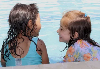 Two toddlers with wet hair and smile at each other on pool ledge during water safety class.