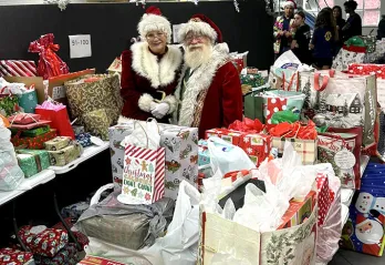 Santa and Mrs. Clause stand around many bags for Giving Tree Gifts.
