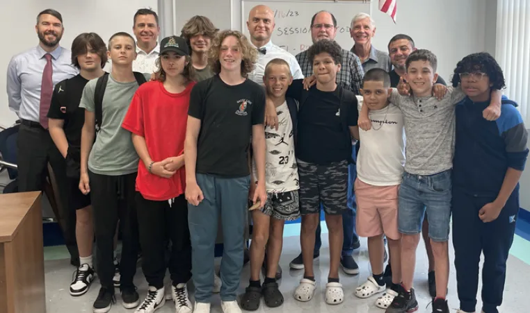 A group of five adults stand behind 10 middle school students, posing for a photo. The setting is a school classroom, a portion of the hanging American flag is shown in the background.