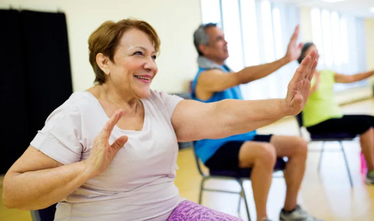 Three seniors holding arms out during chair exercises in fall prevention program. Woman wearing purple pants is in focus, two seniors in the background are out of focus.