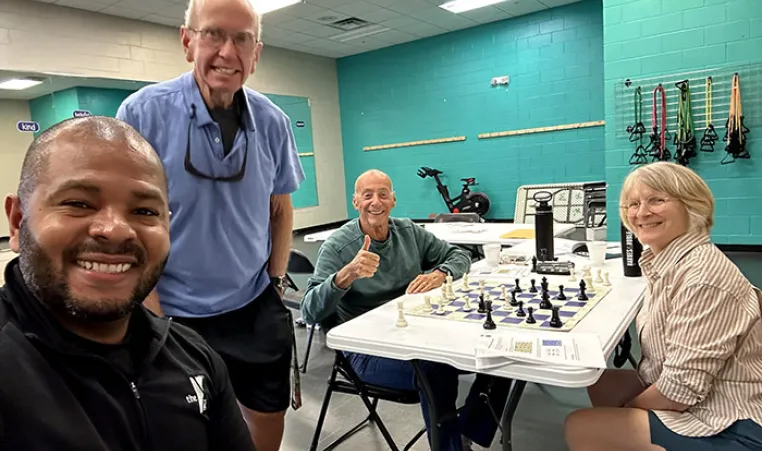 4 members of Chess Club smiling for photo. Two are sitting beside the chess table.