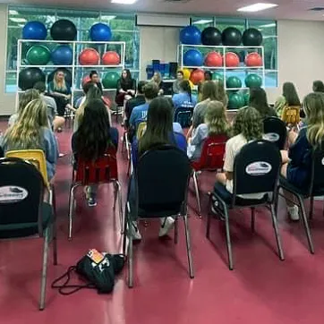 Teens in a large room with red floors, sitting in chairs listening to adult speaker during a meeting.