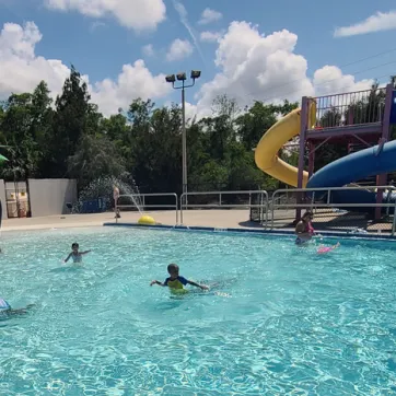 Kids playing in outdoor zero-entry pool with waterslides in the background. Sunny day with puffy white clouds and tall evergreen trees in background scenery.