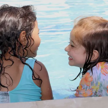 Two toddlers with wet hair and smile at each other on pool ledge during water safety class.