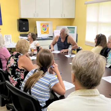 A group of nine adults sitting at a table, listening to a woman standing, teaching the class. Background: classroom walls are yellow.