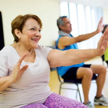 Three seniors holding arms out during chair exercises in fall prevention program. Woman wearing purple pants is in focus, two seniors in the background are out of focus.