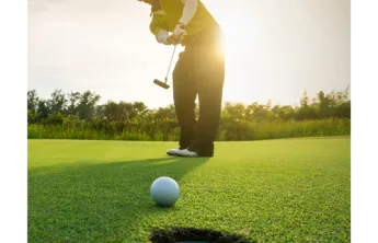 Foreground: golf ball rolling into hole. Background: man in golf attire putting at sunrise. Trees behind him.