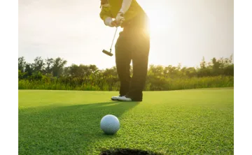 Foreground: golf ball rolling into hole. Background: man in golf attire putting at sunrise. Trees behind him.
